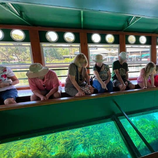 A group of people sitting inside a Glass Bottom Boat, leaning to see the eel grass and other underwater things through the glass in-layed in the center of the boat at Silver Springs State Park in Florida.