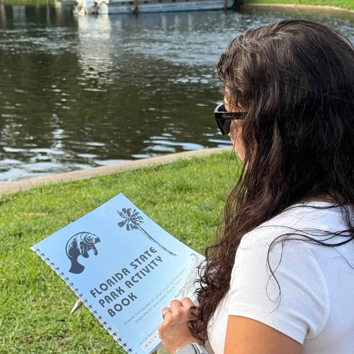 Woman reading a Florida State Park Activity Book while sitting beside a calm river.