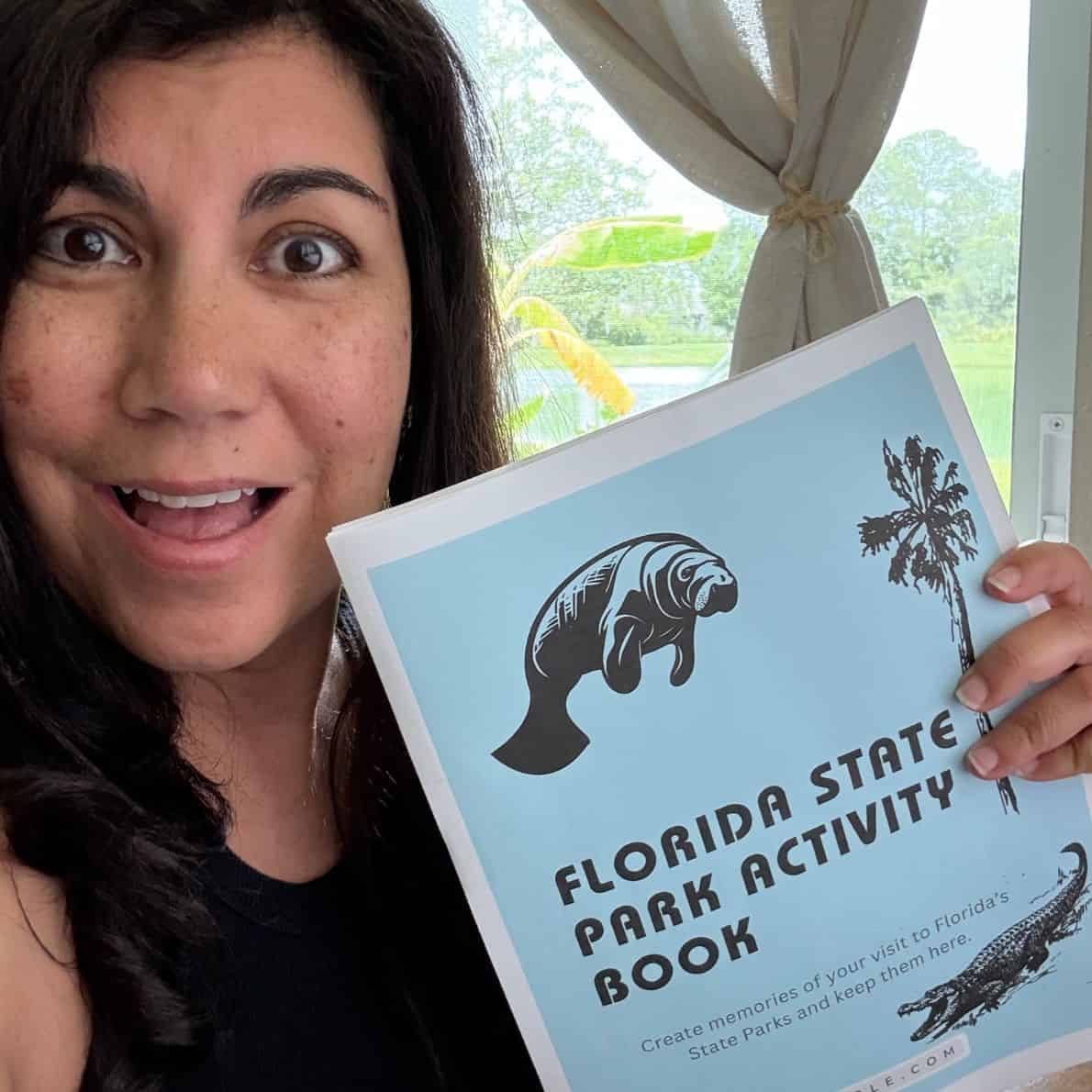 Smiling woman holding a Florida State Park Activity Book with a window and trees in the background.