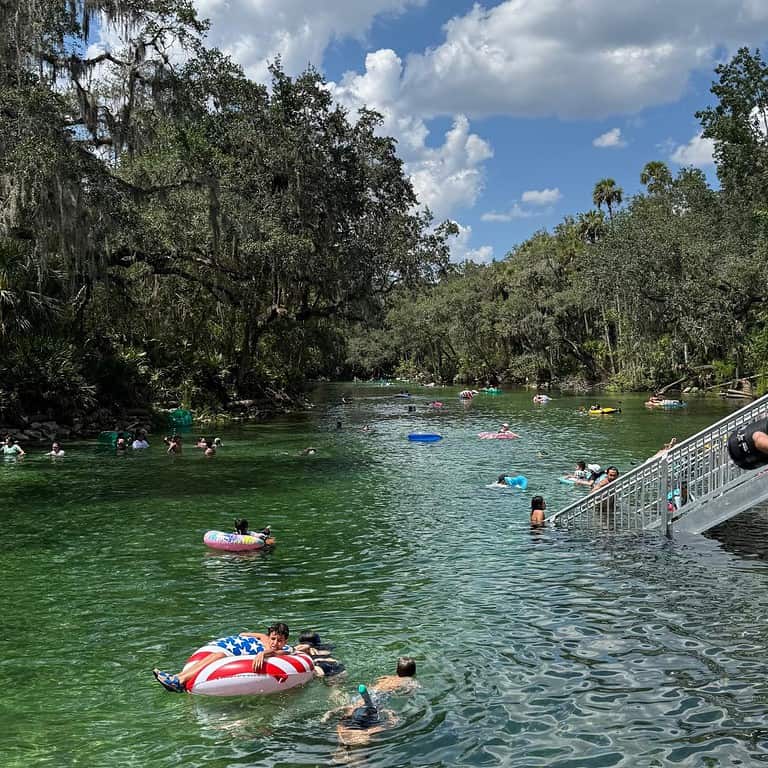 People swimming and floating on tubes in the clear spring run at Blue Spring State Park, with trees and a stair entry visible.