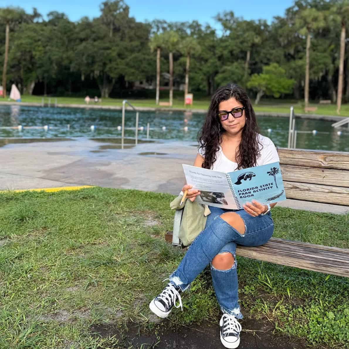 Woman sitting on a bench at De Leon Springs State Park using the Florida State Park Activity Book with the spring pool in the background.