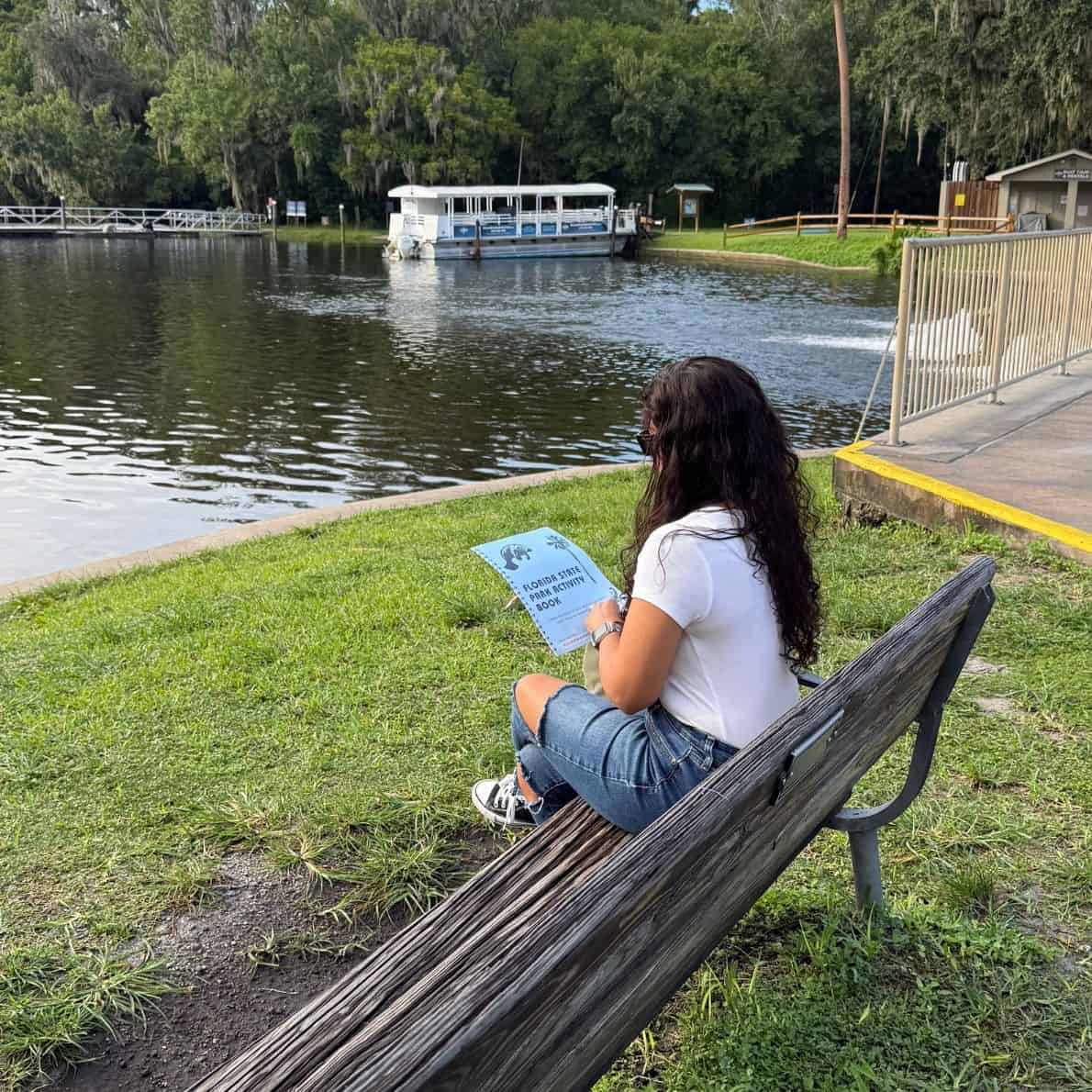 Woman sitting on a bench near the water at De Leon Springs State Park, holding a Florida State Park Activity Book.