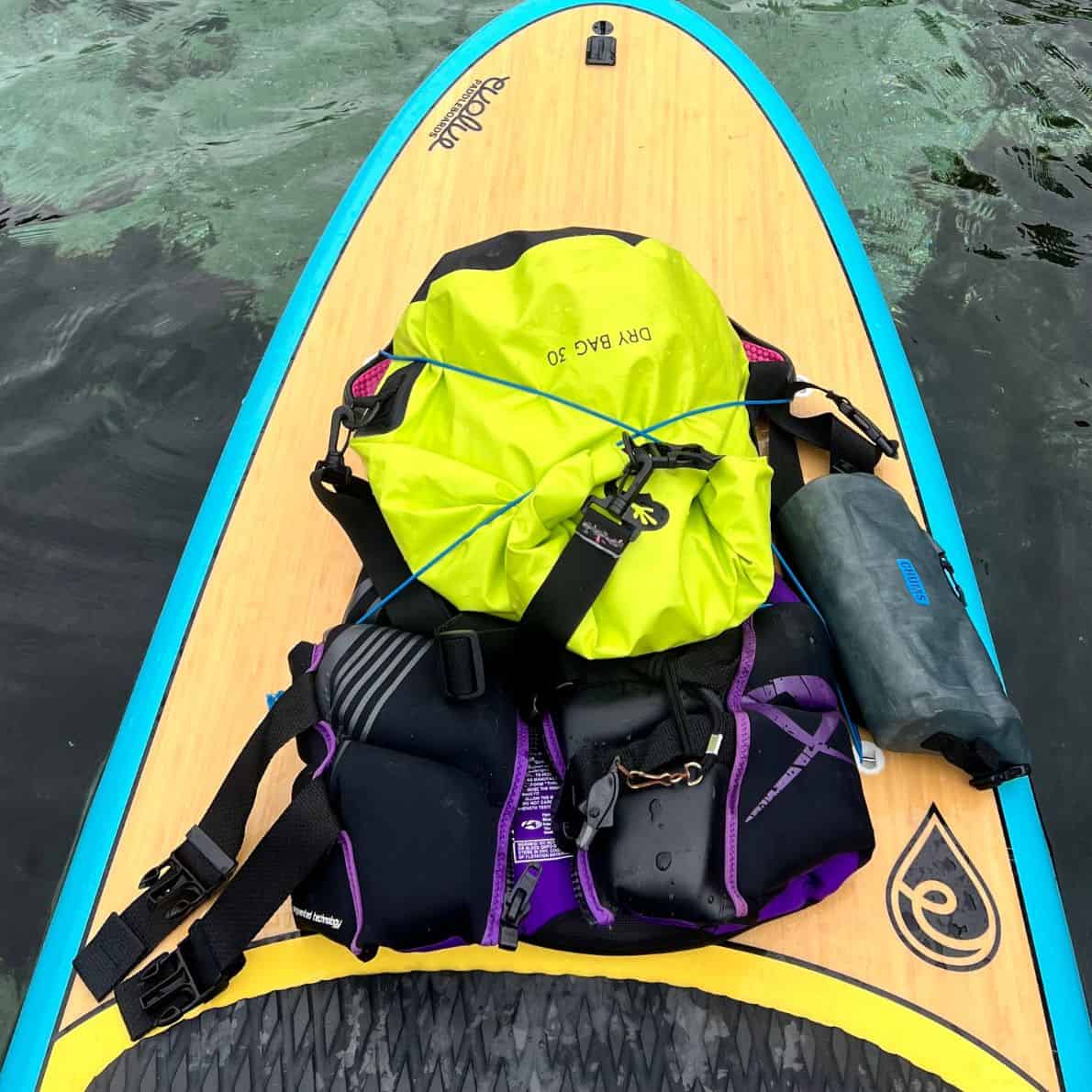 Paddleboard deck loaded with a bright yellow dry bag, gray dray bag, life jacket, and other gear secured with bungee straps.