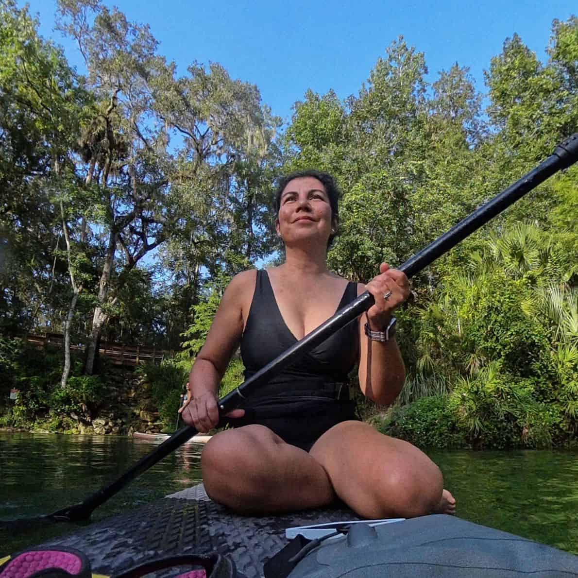 Woman sitting cross-legged on a paddleboard holding a paddle at Blue Spring State Park with green trees in the background.