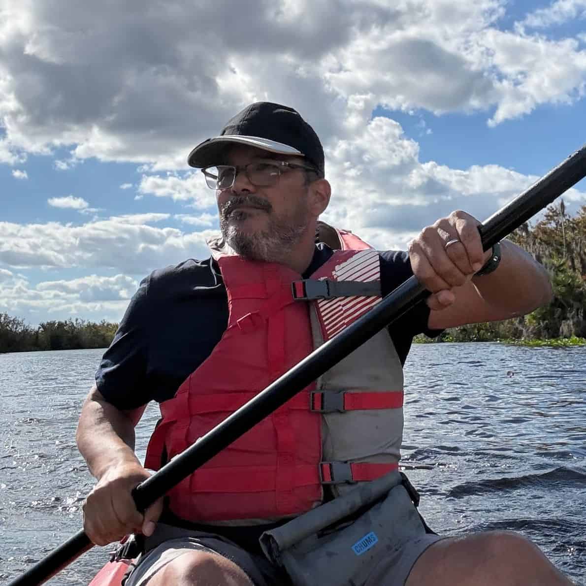 Man wearing a red life jacket kayaking under blue skies at De Leon Springs State Park.
