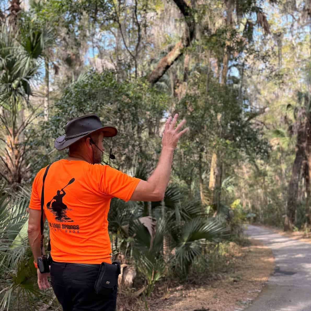 Park guide wearing an orange shirt gestures while leading a guided hike on a paved trail at De Leon Springs State Park.