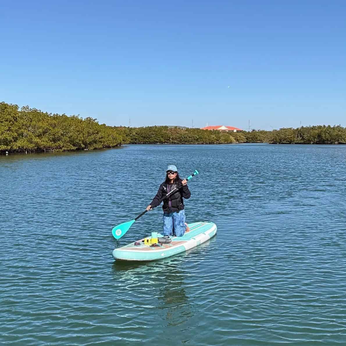 Woman kneeling on a Niphean paddleboard in open water at Callalisa Creek, red-roof building in the distance.