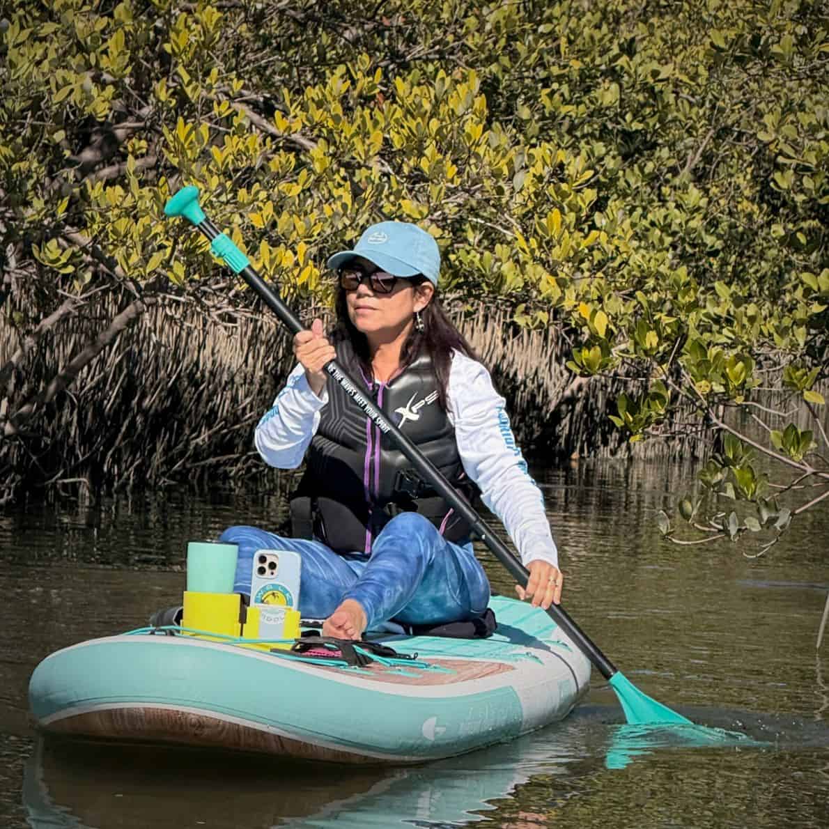 Woman paddling seated on a teal Niphean paddleboard beside mangroves at Callalisa Creek, New Smyrna Beach.