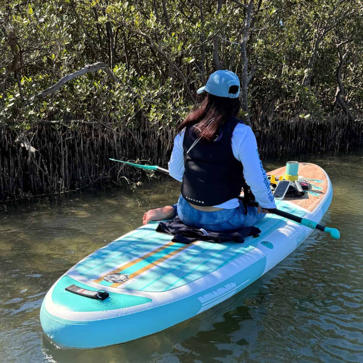 Woman sitting on a teal Niphean paddleboard facing mangroves in shallow water at Callalisa Creek.