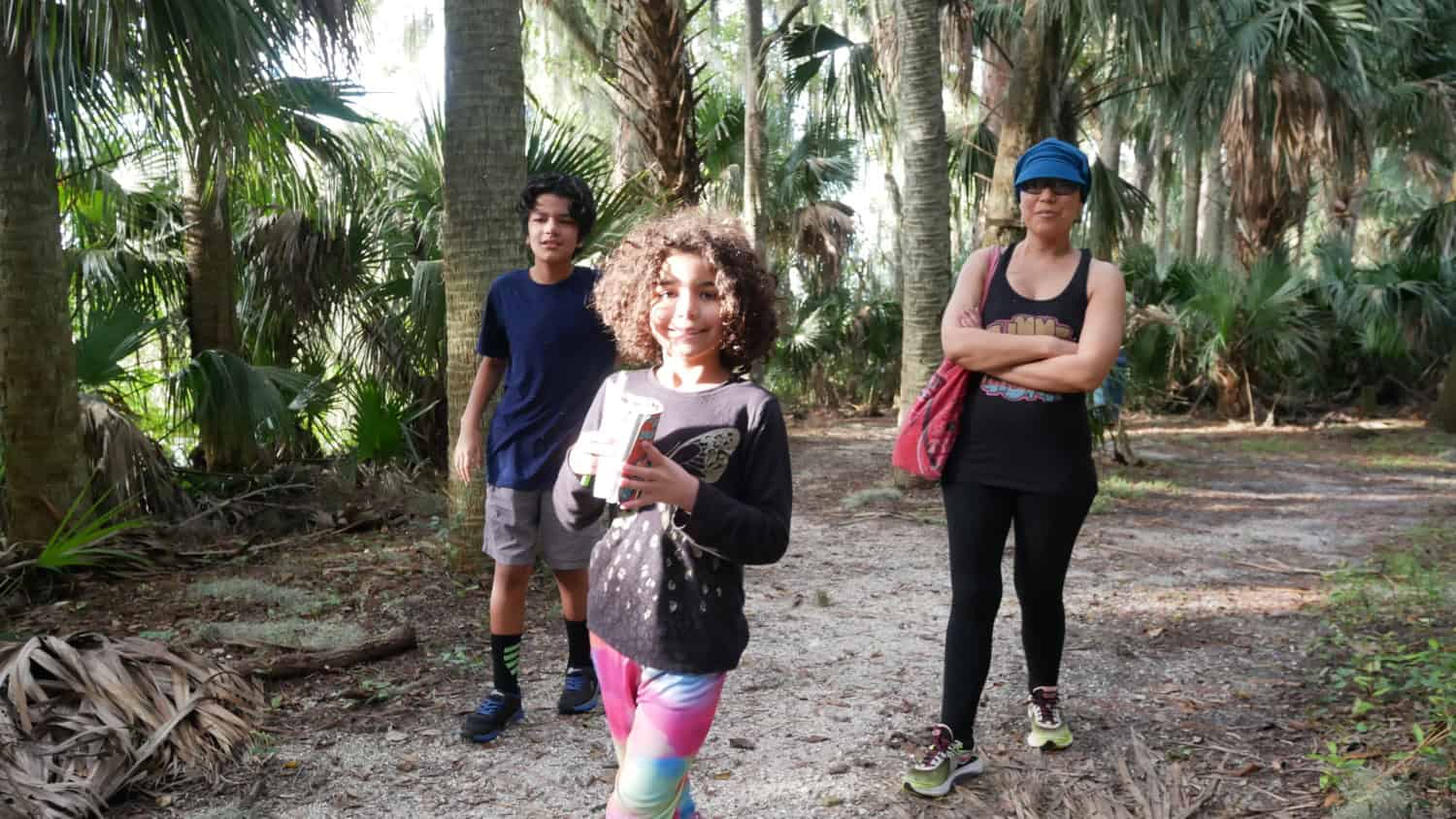 Woman and two children walking along a shaded trail surrounded by palms and trees.