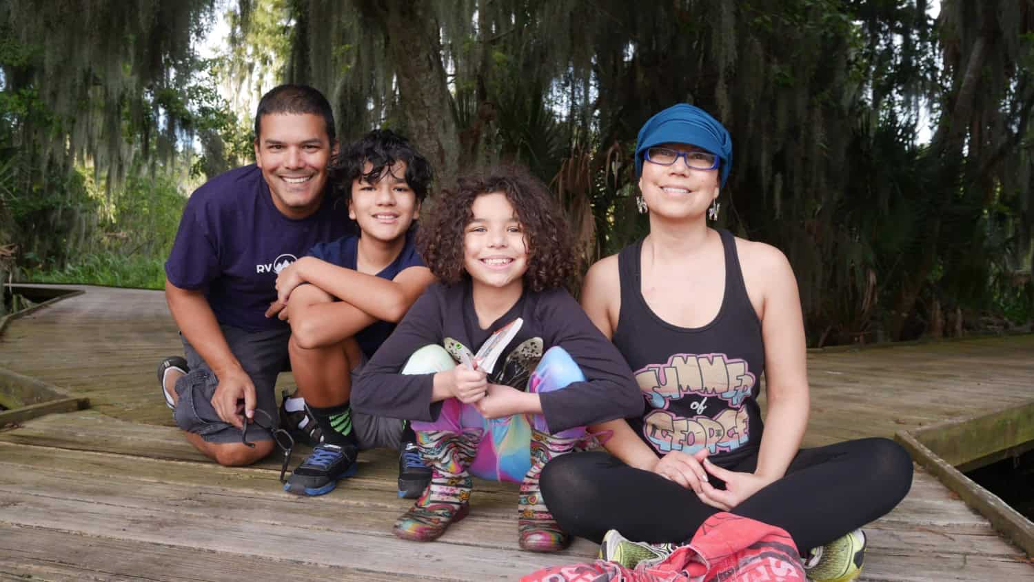 Family of four sitting together on a wooden boardwalk surrounded by trees and Spanish moss.